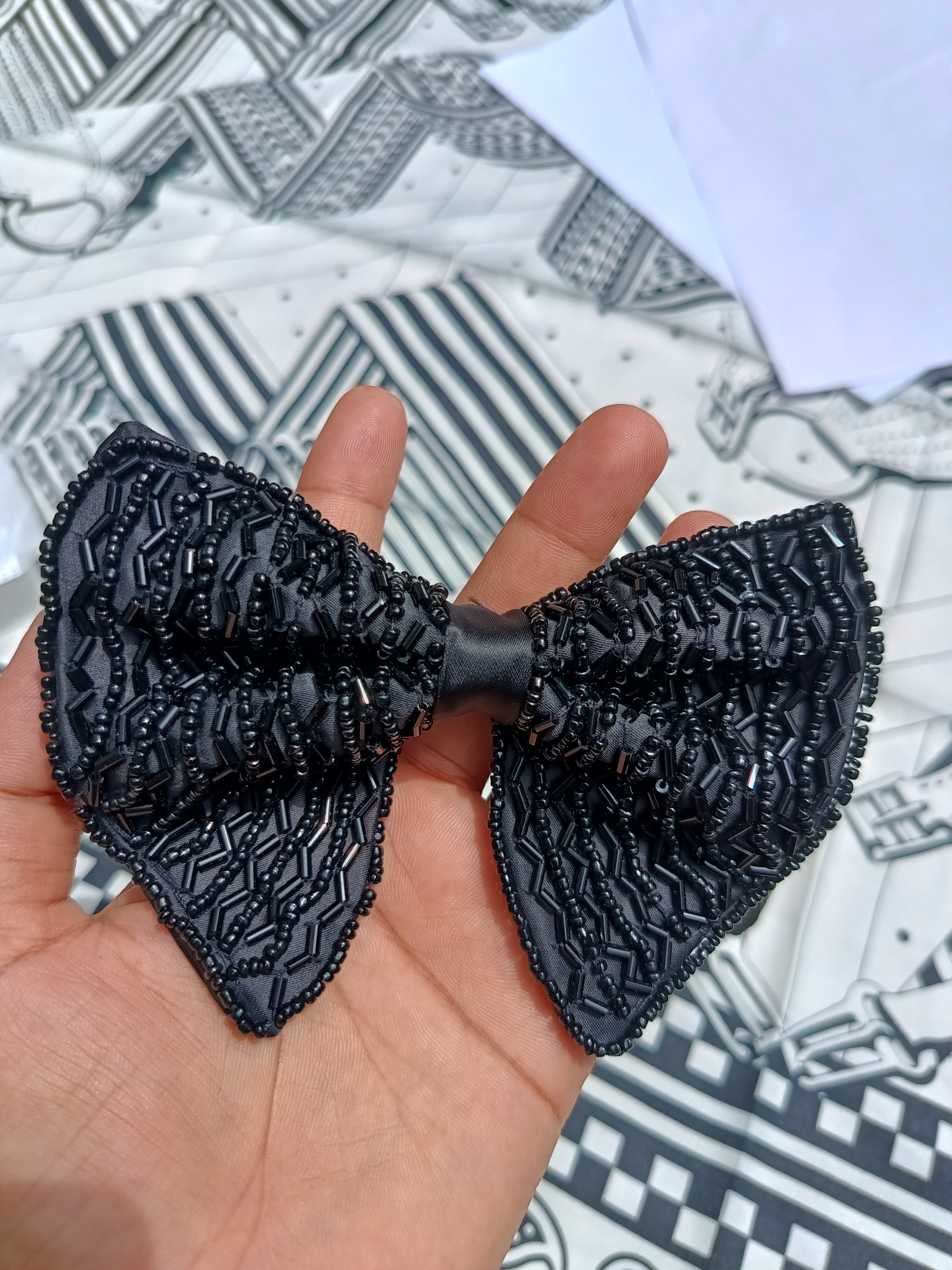 A black textured bow tie with beaded details, held in a person's hand against a patterned black and white fabric background.