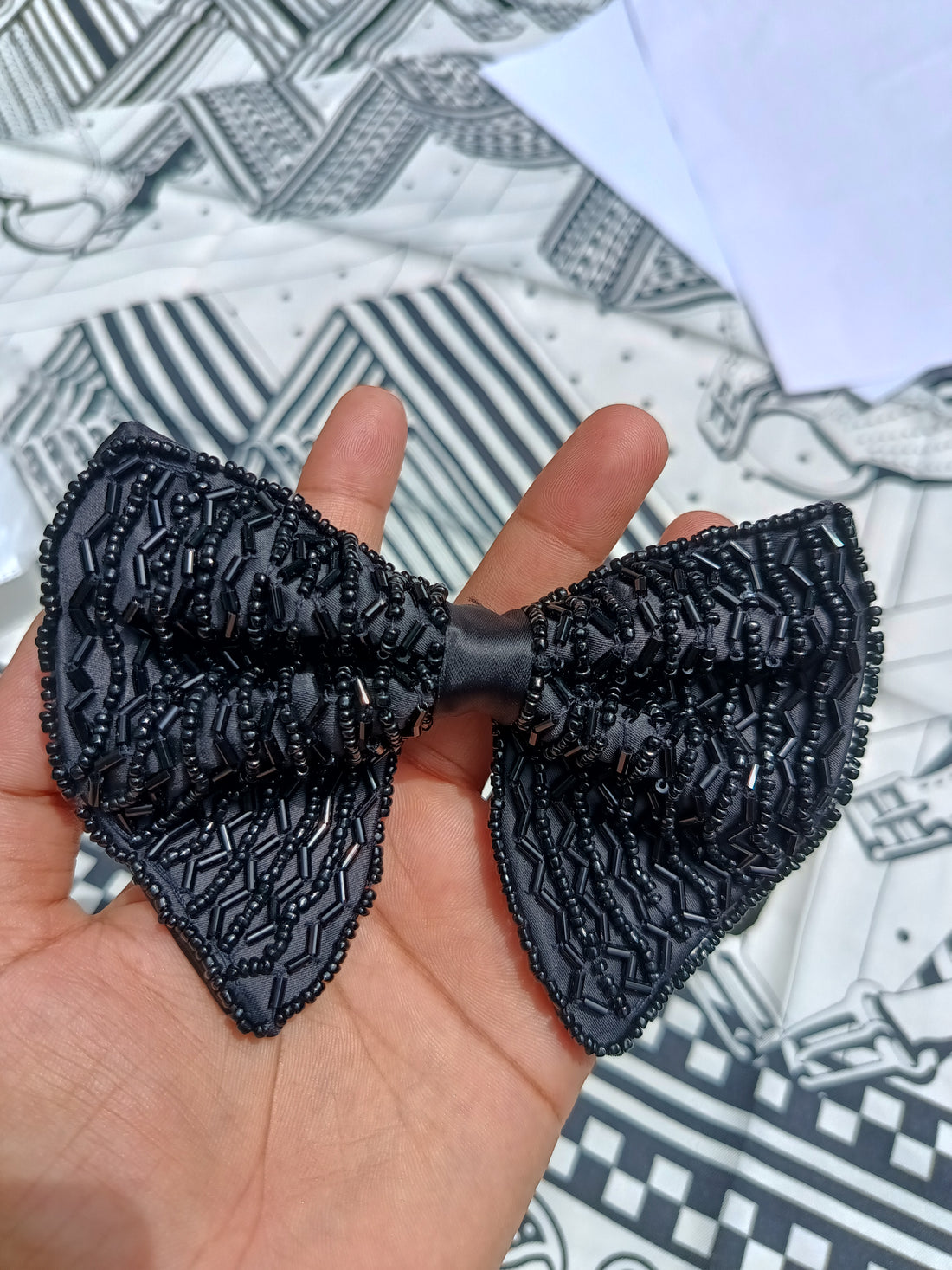 A black textured bow tie with beaded details, held in a person's hand against a patterned black and white fabric background.