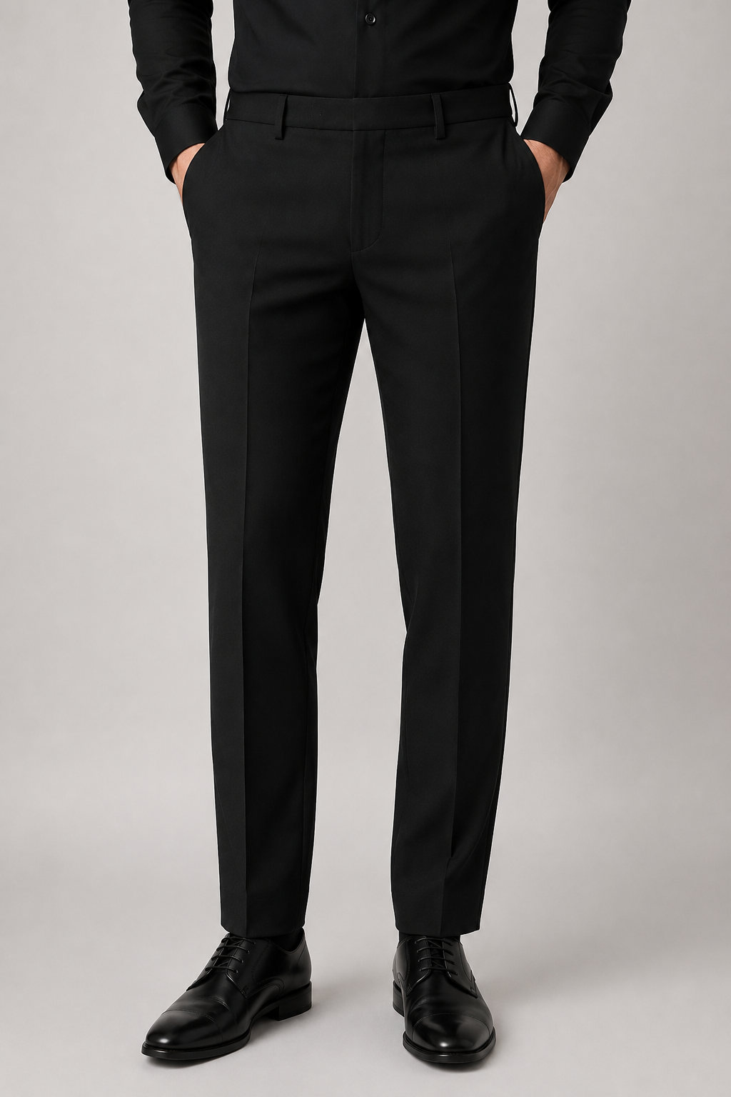 A person wearing black tailored trousers with a black shirt and black dress shoes, shown from the waist down against a light gray studio background.
