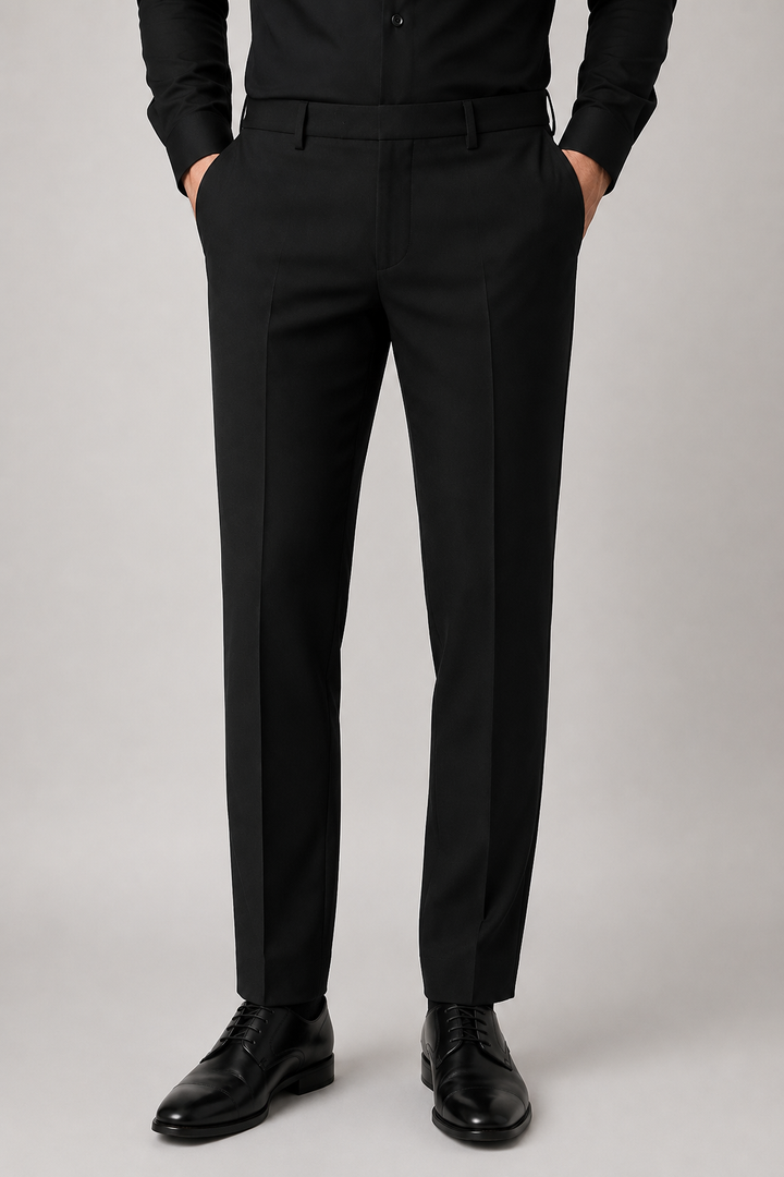 A person wearing black tailored trousers with a black shirt and black dress shoes, shown from the waist down against a light gray studio background.