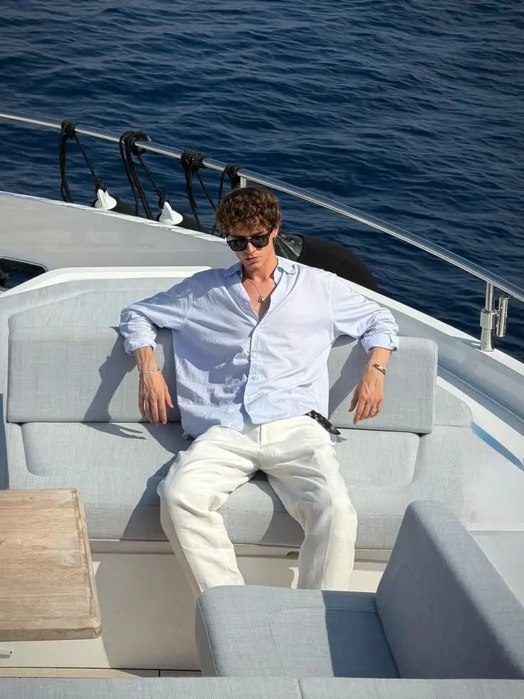 A man wearing a light blue button-down shirt and white trousers relaxes on a light gray cushioned boat seat with blue ocean water in the background.