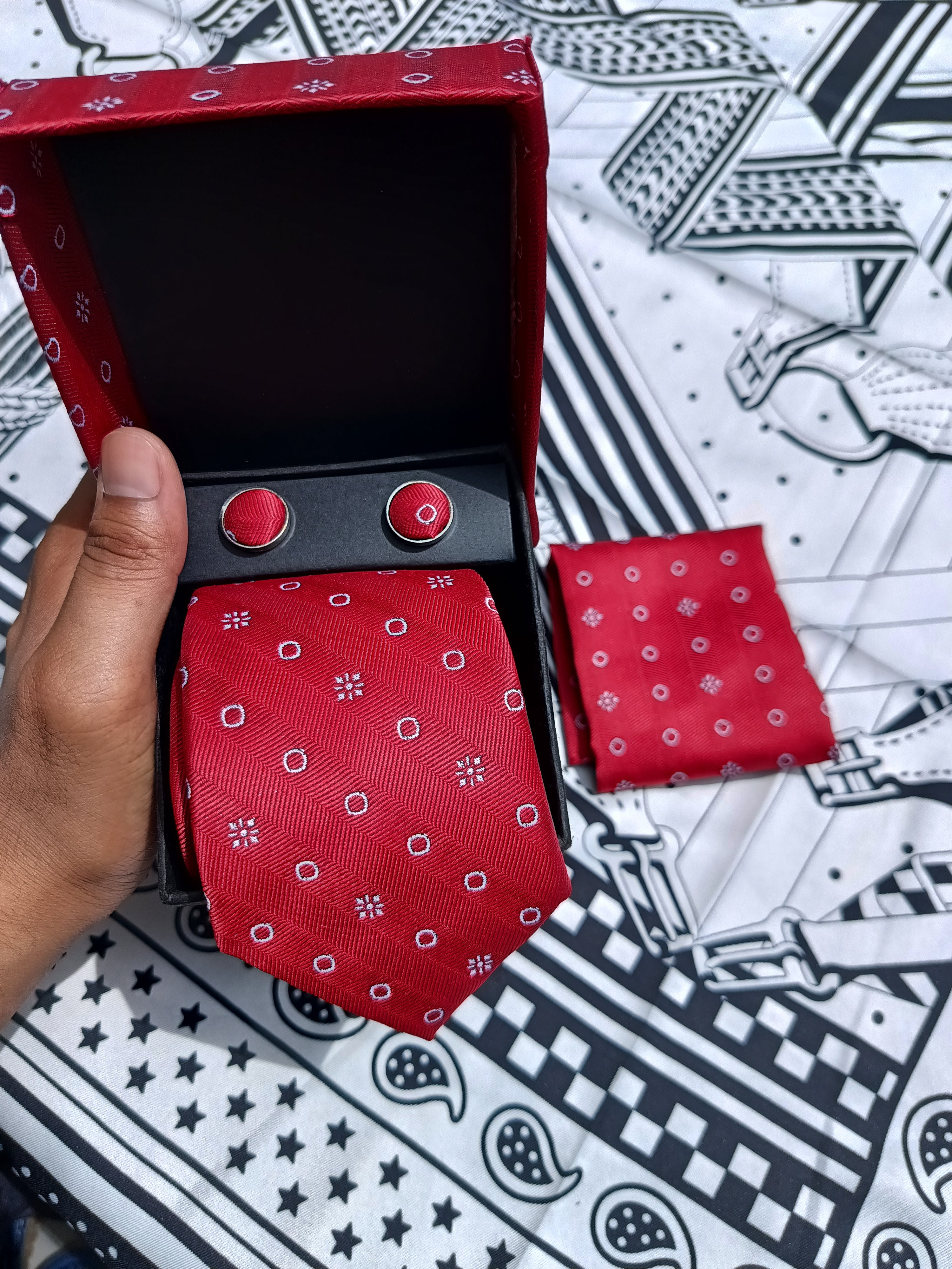 A red necktie with a small white geometric pattern is displayed in an open matching red gift box, alongside a folded matching pocket square and two round red cufflinks on a black insert. A hand is holding the box, and the background is a black-and-white patterned fabric.
