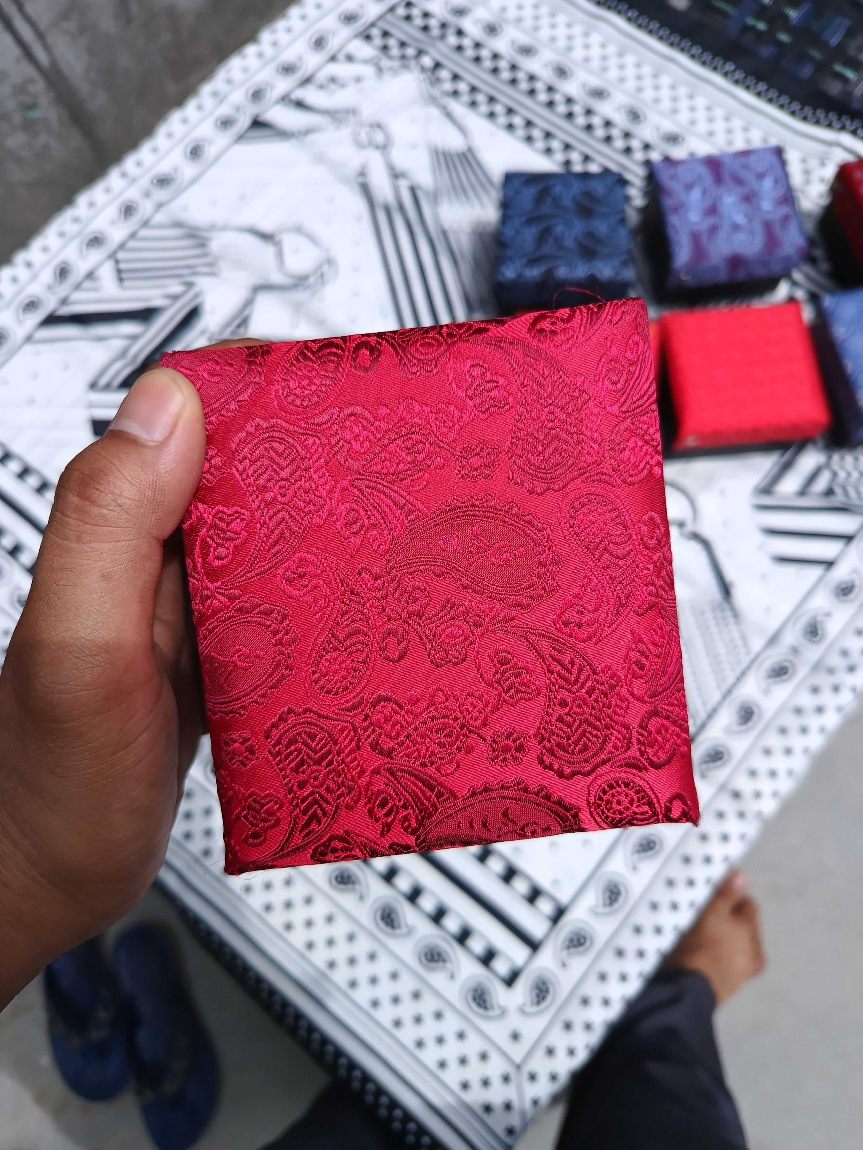A hand holds a small square red pouch or envelope with an embossed paisley pattern, photographed over a patterned tablecloth with other small colorful boxes in the background.