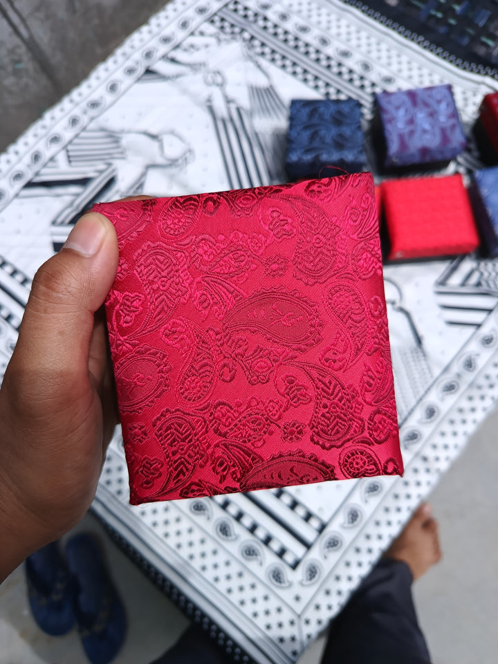 A hand holds a small square red pouch or envelope with an embossed paisley pattern, photographed over a patterned tablecloth with other small colorful boxes in the background.