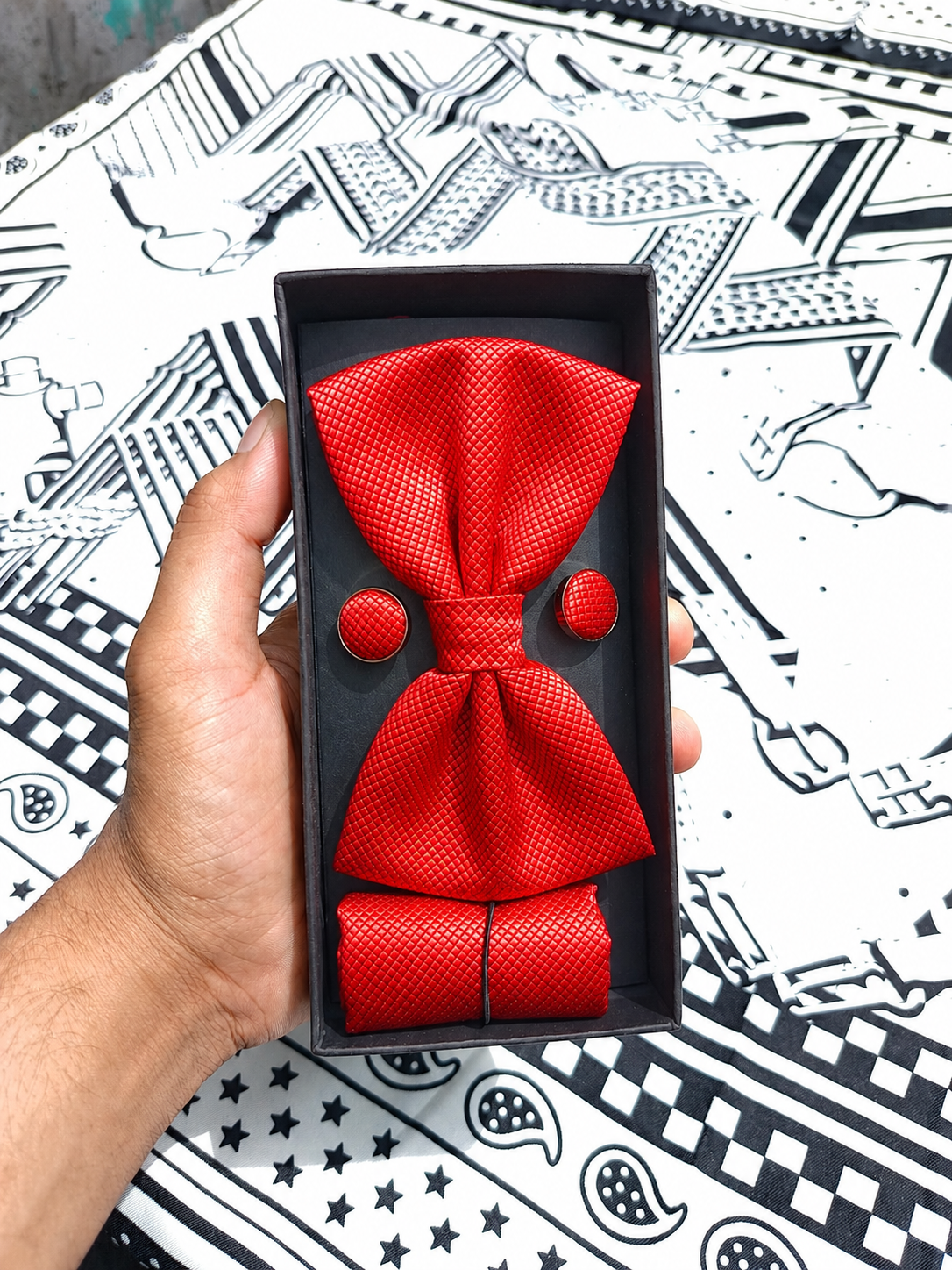A red textured three-piece men's accessory set, including a bow tie, matching pocket square, and two round cufflinks, presented in a black gift box and held by a hand over a black and white patterned surface.