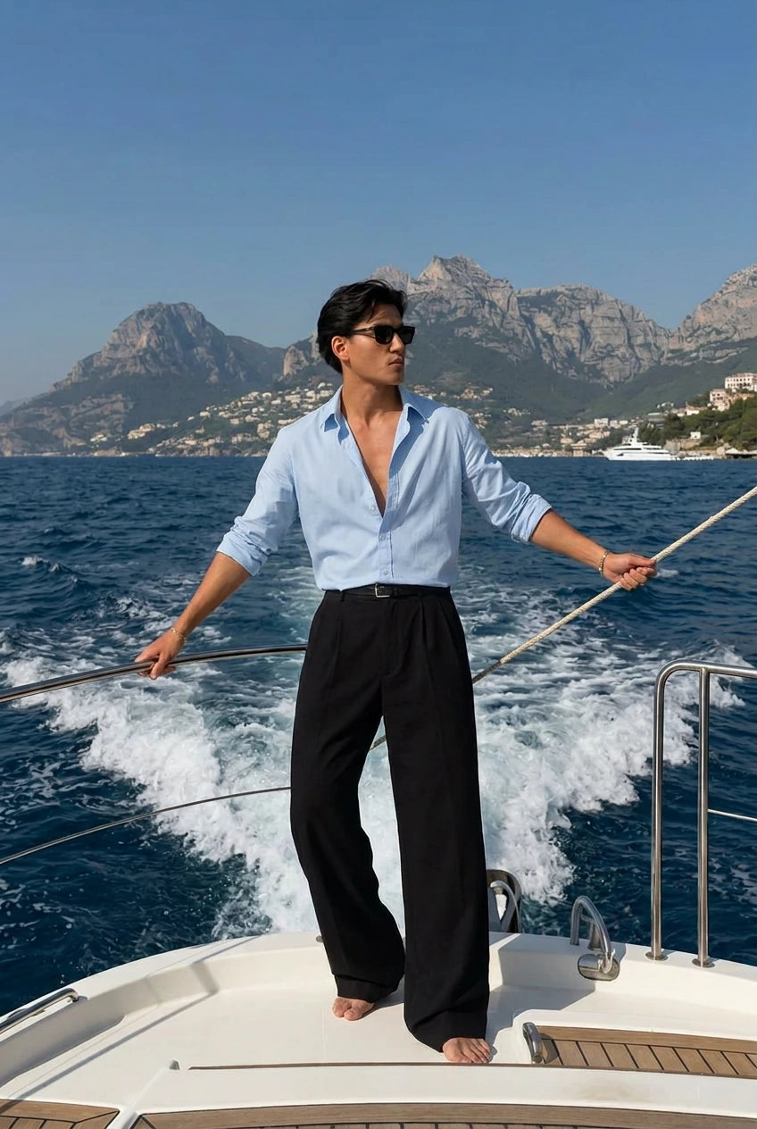 A man wearing a light blue open-collar dress shirt and black trousers stands on the bow of a yacht, holding the tiller with the sea and mountains in the background.
