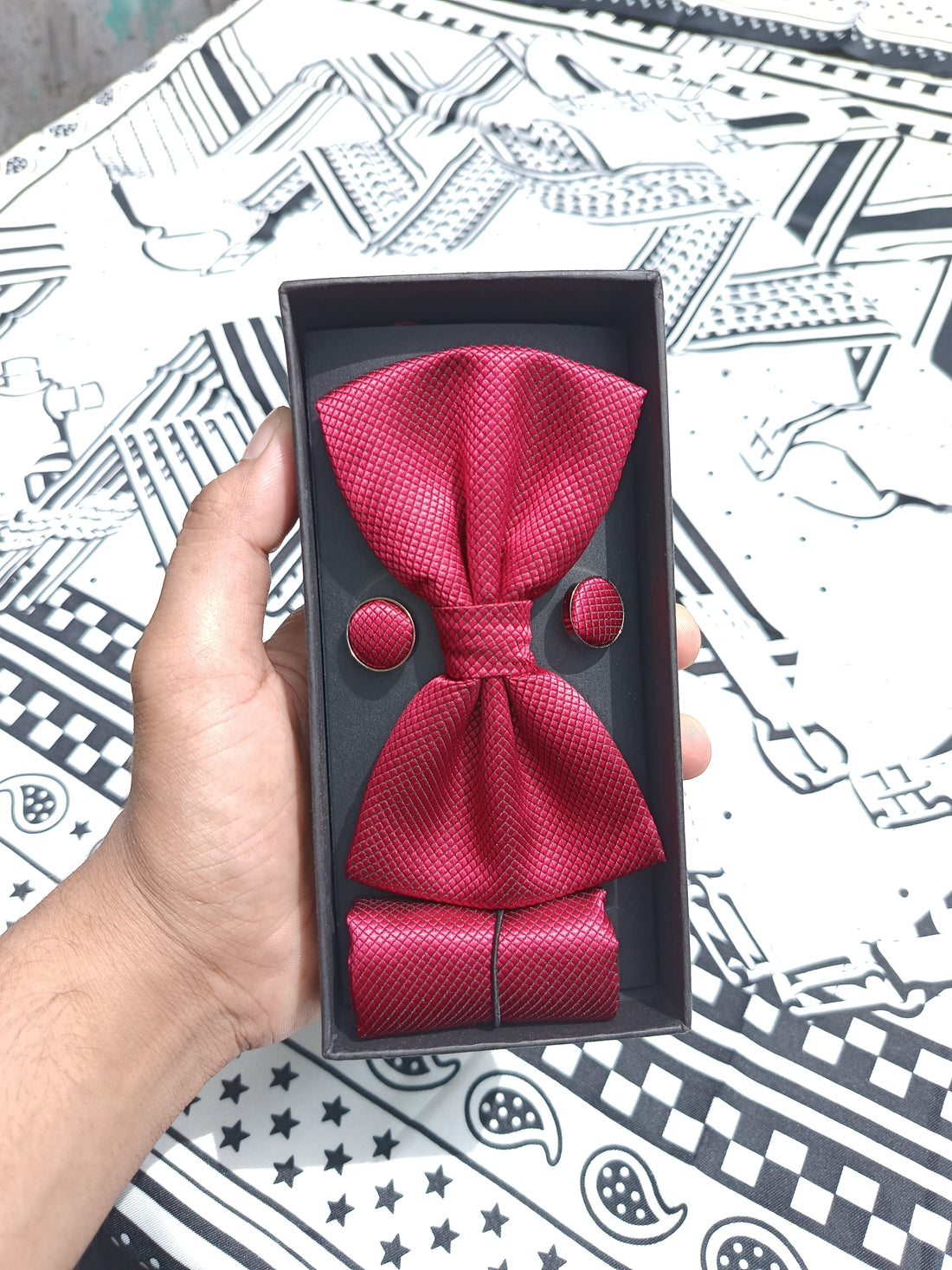 A dark red textured bow tie set with matching buttons and a matching necktie, presented in a black gift box held by a hand over a black and white patterned fabric background.